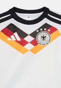 Λευκή ποδοσφαιρική φανέλα με διαγώνιες μαύρες, κόκκινες και κίτρινες ρίγες, διαθέτει στρογγυλή λ�αιμόκοψη, τρεις μαύρες ρίγες στους ώμους και έμβλημα.