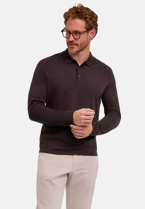 Mann mit lockigem Haar, der eine Brille, ein dunkelbraunes langärmliges Poloshirt und hellbeige Hose trägt, steht und richtet einen Ärmel.
