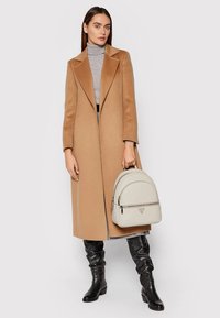 Lungo cappotto di cammello con colletto a punte, dolcevita grigia, stivali neri fino al ginocchio e uno zaino in pelle grigia chiara con zip e logo.