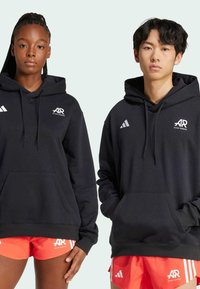 Crne dukserice s kapuljačom i prednjim džepovima, s bijelim Adidas i AR logotipima, uparenim s svijetlo narančastim šortsevima. Glatka tekstura pamuka.