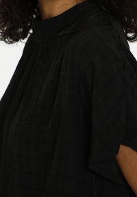 Haut noir avec un col montant, des manches courtes amples, et un tissu texturé présentant de subtiles lignes horizontales.