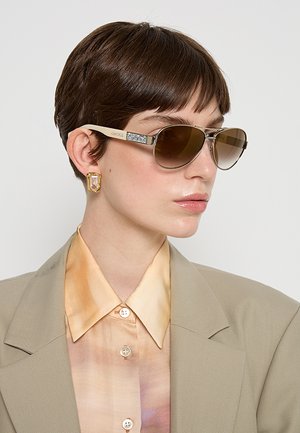 Donna con capelli corti castani che indossa occhiali da sole aviator con lenti marroni, orecchini geometrici dorati, camicia beige pastello con bottoni e blazer marrone chiaro.