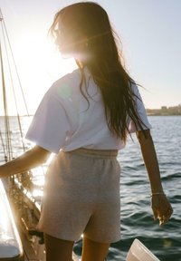 T-shirt bianco oversize e pantaloni corti grigi chiari con cintura elastica, visti da dietro vicino a una barca a vela e all'acqua, con il sole dietro.