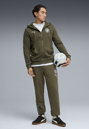 Mężczyzna w oliwkowo-zielonym dresie Puma trzyma piłkę nożną, stoi z nogami lekko rozstawionymi, patrząc na bok na szarym tle.