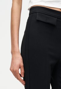 Γυναικείο χέρι που ακουμπάει στο πλάι, φοράει μαύρο παντελόνι με ψηλή μέση και διακοσμητικό καπάκι, με λευκό τοπ να φαίνεται πάνω από τη μέση.