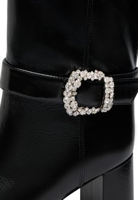 Stivaletto in pelle nera con fibbia quadrata decorata da strass, texture liscia e tacco chunky. I dettagli includono una finitura lucida e cuciture.