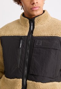 Fleecegefütterte Jacke mit einem oberen Abschnitt in Beige und einem unteren Abschnitt in Schwarz. Sie verfügt über zwei mit Reißverschluss verschlossene Taschen und ein strukturiertes schwarzes Patch.