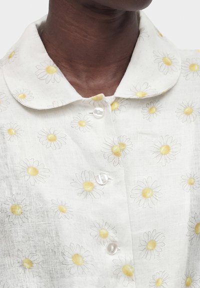 Λευκό πουκάμισο με γιακά και τύπωμα μαργαρίτας με κίτρινο κέντρο και τρία μαργαριταρένια κουμπιά, φορεμένο από άτομο με σκουρόχρωμο δέρμα και ορατό λαιμό.