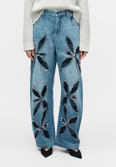 Jean en denim bleu clair à jambes larges avec de grandes appliques florales noires et bords effilochés, porté avec un pull gris et des chaussures noires.