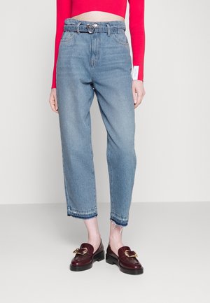 Nainen pukeutuneena vaaleansinisiin korkeavyötäröisiin lyhennettyihin farkkuihin, joissa on raakasaumat, viininpunaisiin loafer-kenkiin kultaisella soljella ja punaiseen pitkähihainen lyhyt toppiin.