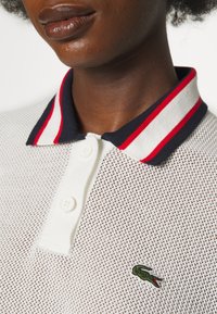 Gros plan d’une personne portant un polo en maille blanche avec un col rayé bleu marine, rouge et blanc, et un logo crocodile vert sur la poitrine.