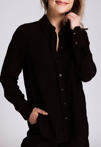 Schwarzes Cordhemd mit Knöpfen, ausgestattet mit Kragen, langen Ärmeln und einer Tasche auf Taillenhöhe. Texturiertes Gewebe mit vertikalen Linien.