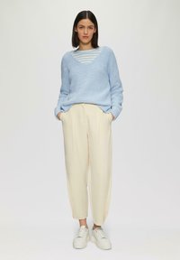 Hellblauer gestrickter Pullover mit V-Ausschnitt, kombiniert mit cremefarbenen, schmal geschnittenen Hosen und weißen Plateausneakern. Gestreiftes Hemd darunter.
