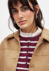 Tansuede-Jacke mit gesteppten Schulterdetails über einem bordeauxfarbenen gestreiften Hemd mit Knopfverschluss und einem weißen Rollkragenpullover darunter.