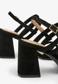 Sorte ruskinds hælede sandaler har en bred blokhælkonstruktion, krydsede stropper og en gylden spænde detaljer, der fremviser en struktureret overflade.