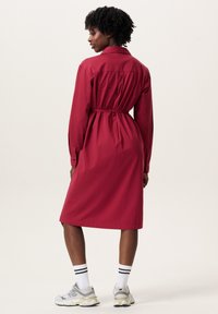 Rotes Hemdkleid mit langen Ärmeln, tailliertem Schnitt und mittellangem Saum. Kombiniert mit weißen Socken und grauen Turnschuhen. Glatte Stofftextur.