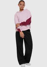 Hoodie in roze en bordeaux, gemaakt van zacht materiaal. Heeft lange mouwen, een relaxte pasvorm en geribde manchetten. Gemaakt in combinatie met zwarte wijde broek.