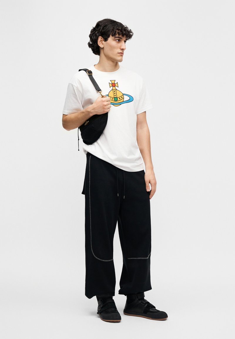 Hvid bomuld T-shirt med et farverigt grafisk design, kombineret med løse sorte bukser og sorte sko, samt en lille sort taske.