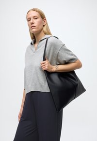 Svart läderaxelväska med en mjuk, strukturerad design. Modellen bär en gråstickad tröja och mörka byxor, vilket visar på väskans mångsidighet.