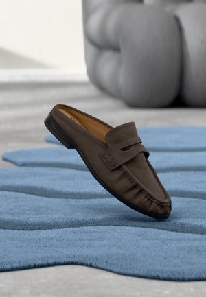Mocassin en daim marron reposant sur un tapis texturé bleu avec un grand objet sculptural gris flou en arrière-plan.