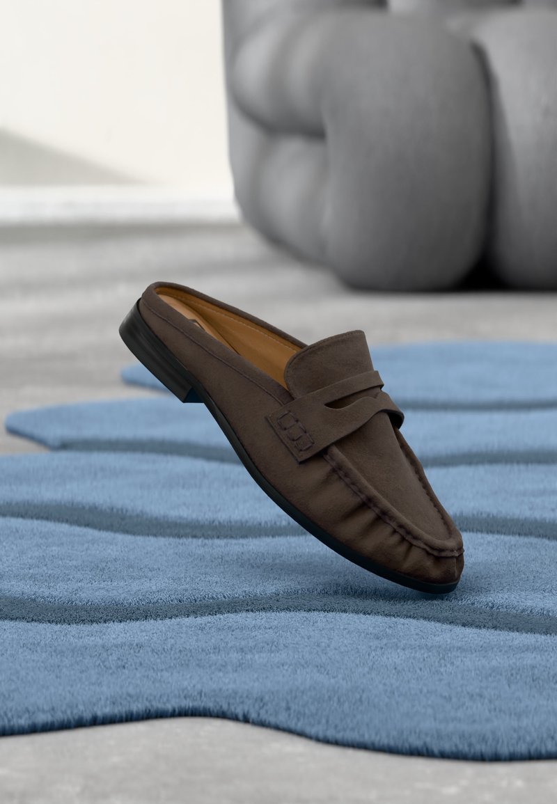 Mocassin en daim marron reposant sur un tapis texturé bleu avec un grand objet sculptural gris flou en arrière-plan.
