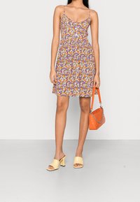 Blumenkleid mit lila, orange und weißen Mustern, tailliertem Oberteil und A-Linien-Rock. Kombiniert mit einer orangefarbenen Handtasche und gelben Blockabsatz-Pantoletten.