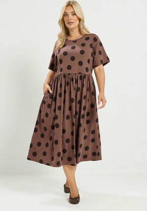 Femme portant une robe midi marron à pois noirs, manches courtes, poches, et des chaussures plates marron, debout devant un fond uni.