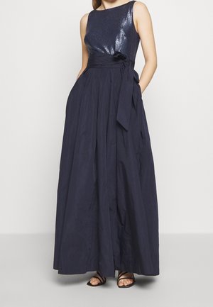 Vrouw draagt een mouwloze marineblauwe jurk met een glanzende top, een brede rok, een gestrikte tailleriem en zwarte sandalen met bandjes.