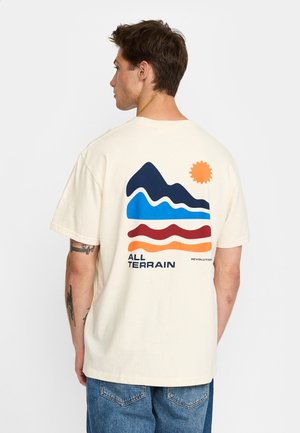 Hombre con camiseta color blanco roto con gráfico colorido de montaña y sol y texto "ALL TERRAIN", de espaldas, mostrando la parte trasera de la camiseta.
