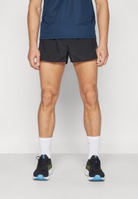 Schwarze Sportshorts, kombiniert mit einem blauen Sportshirt, weißen Socken und schwarzen Laufschuhen mit grünen und blauen Akzenten.