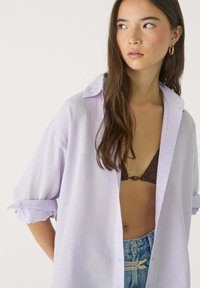 Jasnoliliowa lniana koszula z podwiniętymi rękawami, luźny krój, zapinana na guziki, noszona na ciemnobrązowym bikini.