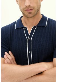 Marineblauwe textuur polo-shirt met korte mouwen en een witte afgewerkte kraag, voorzien van een knoopsluiting en een kenmerkend verticale ribbelpatroon.