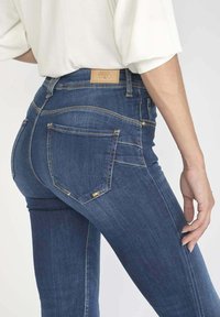 Jean skinny en denim bleu foncé. Présente un matériel doré, deux poches arrière et des coutures détaillées. Design taille haute avec une coupe classique.