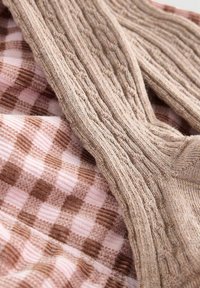 Maglione lavorato a maglia color beige con motivi a treccia testurizzati, sovrapposto a un tessuto a quadretti rosa e marrone, caratterizzato da texture lisce e a coste in contrasto.