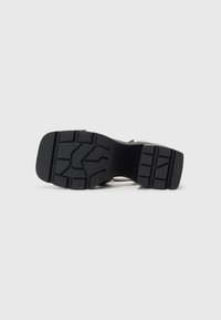 Semelle de chaussure noire avec des motifs de bande de roulement profonds, talon carré et adhérence texturée, vue du dessous sur un fond clair uni.