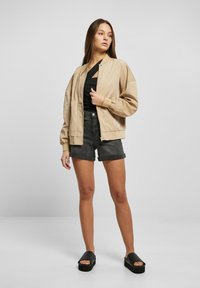 Giacca bomber beige, taglio oversize con chiusura a zip, indossata sopra una maglietta nera e pantaloni corti grigi; abbinata a sandali neri con zeppa.