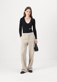 Black long-sleeve bodysuit with a deep V-neck and white logo, paired with beige wide-leg trousers, and black bag. Smooth textures.