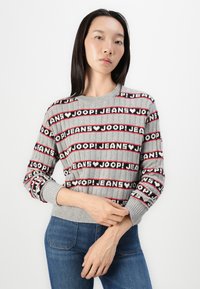 Maglione grigio lavorato a maglia con texture a costine, caratterizzato da un motivo grafico ripetuto in nero, rosso e bianco. Maniche lunghe e scollatura rotonda.