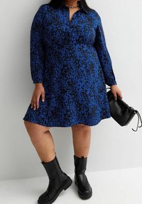 Blaues Kleid mit schwarzem Spritzmuster, langen Ärmeln, Kragen und schwingendem Saum. Kombiniert mit schwarzen Stiefeln mit dicker Sohle und einer kleinen schwarzen Tasche.