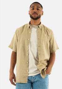 Camicia button-up a maniche corte color beige chiaro con un logo sul petto, indossata sopra una maglietta grigia chiara a girocollo, abbinata a jeans blu.