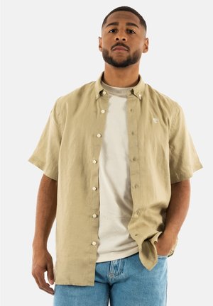 Camicia button-up a maniche corte color beige chiaro con un logo sul petto, indossata sopra una maglietta grigia chiara a girocollo, abbinata a jeans blu.