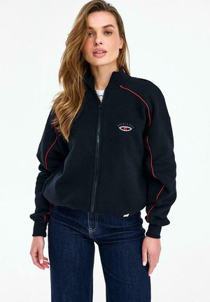 Kobieta nosząca czarną kurtkę na zamek z czerwonymi lamówkami i haftowanym wzorem oka na piersi, w połączeniu z ciemnoniebieskimi jeansami.