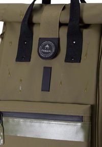 Rucksack aus olivgrünem Stoff mit schwarzen Gurten und Logo-Patch. Verfügt über eine silberne Reißverschlusstasche, Wassertropfen auf der Oberfläche und eine glatte Textur.