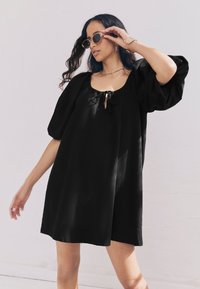 Sort, kortærmet kjole med løs pasform, der har en firkantet halsudskæring og bindedetalje foran. Glat tekstur med et afslappet design.
