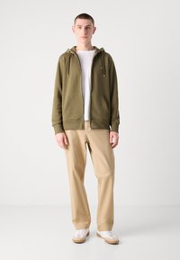 Olijfgroene zip-up hoodie met twee voorkruimels, gedragen over een wit T-shirt, gecombineerd met beige broeken en witte sneakers.