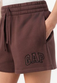Brune bomuldsshorts med elastisk talje, snor og sidelommer; har sort broderet "GAP" logo på forsiden.