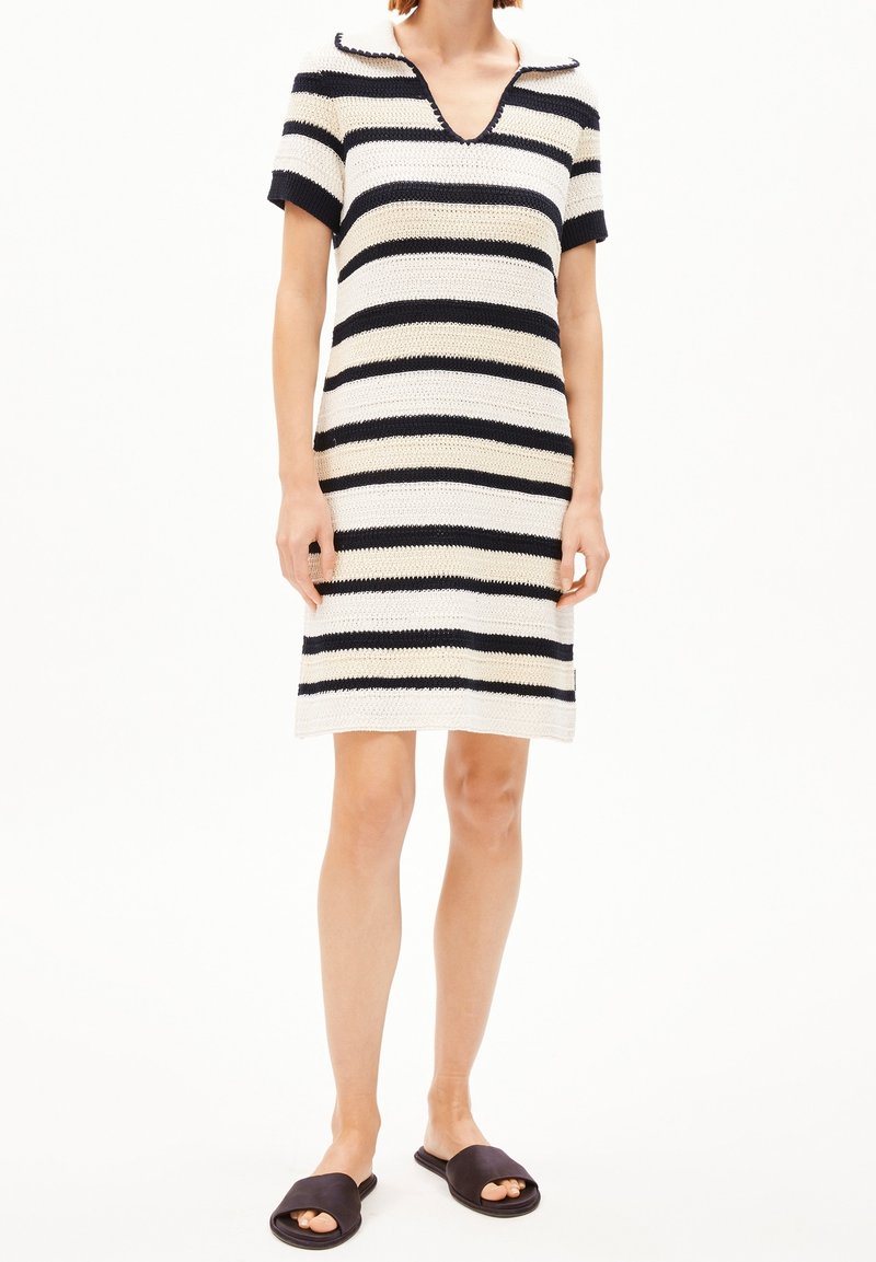 Femme portant une robe en maille rayée aux manches courtes et longueur genou, ainsi que des sandales noires à bouts ouverts, se tenant devant un fond blanc.