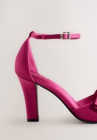 Vaaleanpunainen satiininen korkokenkä nilkkaremillä, jossa rusetti edessä ja paksu korko. Sileä pinta ja eloisa väri.