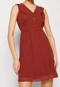 Sukienka bez rękawów w kolorze rdzy z tkaniny w fakturę kropek, z dekoltem w kształcie litery V, zapinana na guziki z przodu oraz z falbanami na ramionach, noszona przez stojącą osobę.