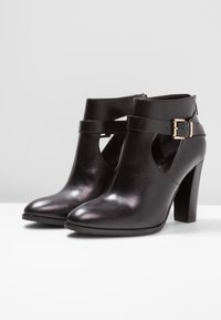 Botines de cuero negro con tacones altos, con un diseño cortado, correa en el tobillo con detalle de hebilla dorada y textura suave y brillante.